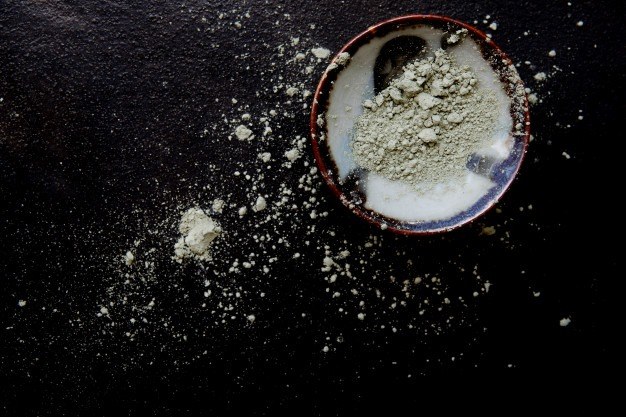 Argile cosmétique verdâtre en poudre dans un bol en céramique sur un fond sombre, poudre répandue autour.