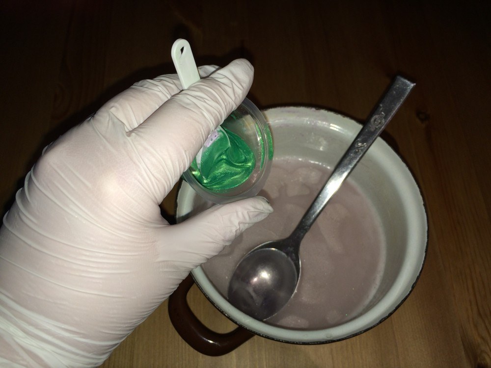 Védőkesztyűs kéz zöld elkevert színezéket tartalmazó edénykét tart a lilára színezett, felolvasztott szappanalapot tartalmazó bögre fölött.