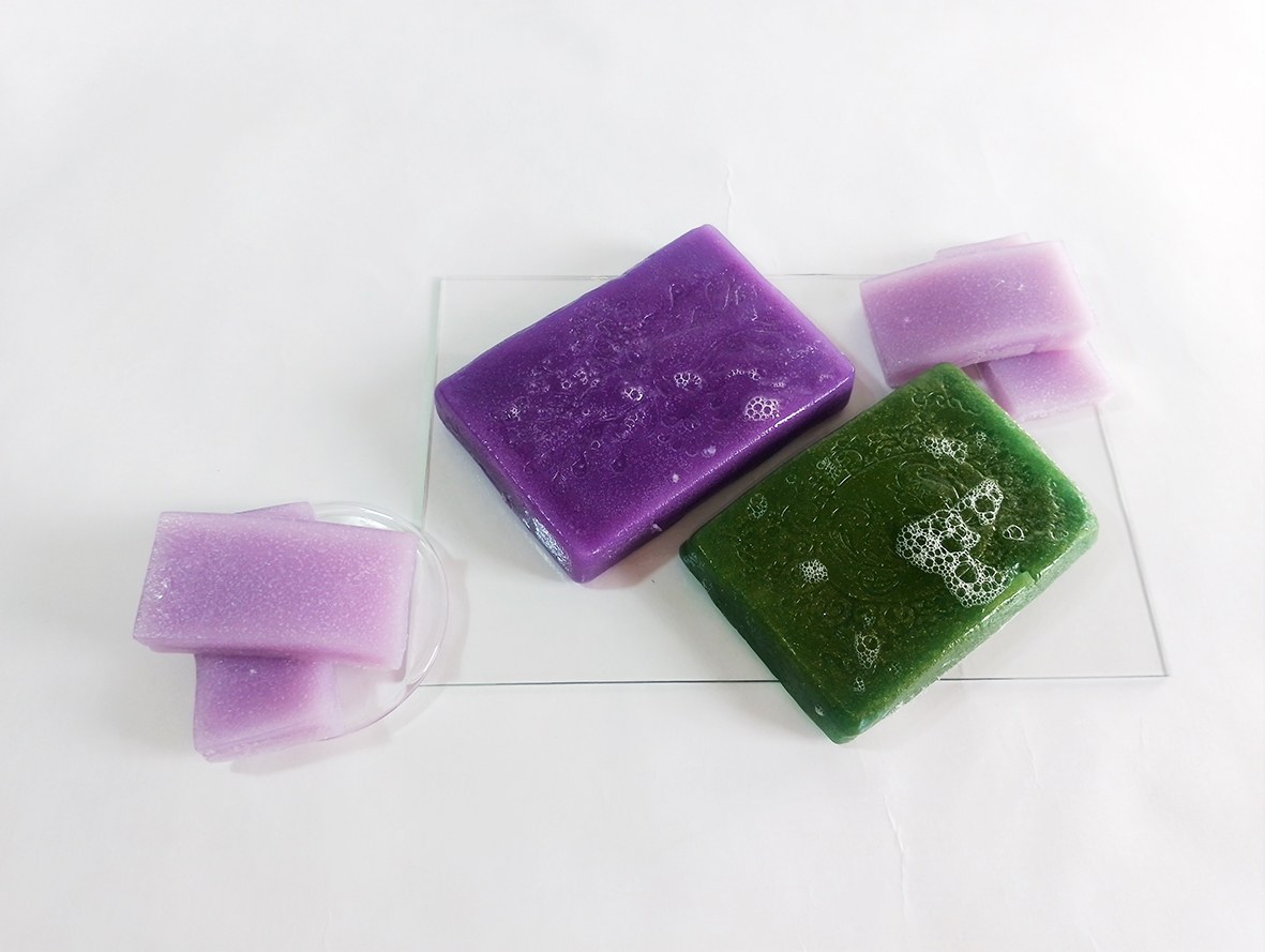 Jelly soap in lavender, purple and green colour on a transparent acrylic pad placed on a white background.