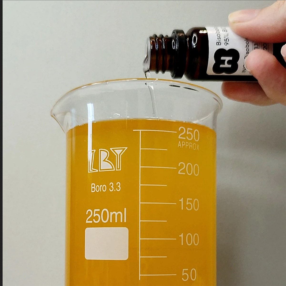 Une main fait tomber le bisabolol goutte à goutte d’un flacon foncé dans un bécher en verre rempli d’un mélange d’huiles orangé.