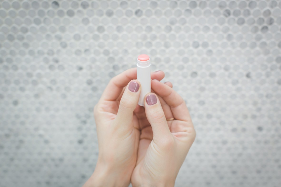 Una mujer sostiene el bálsamo labial de cuidado en las manos sobre un fondo gris claro.