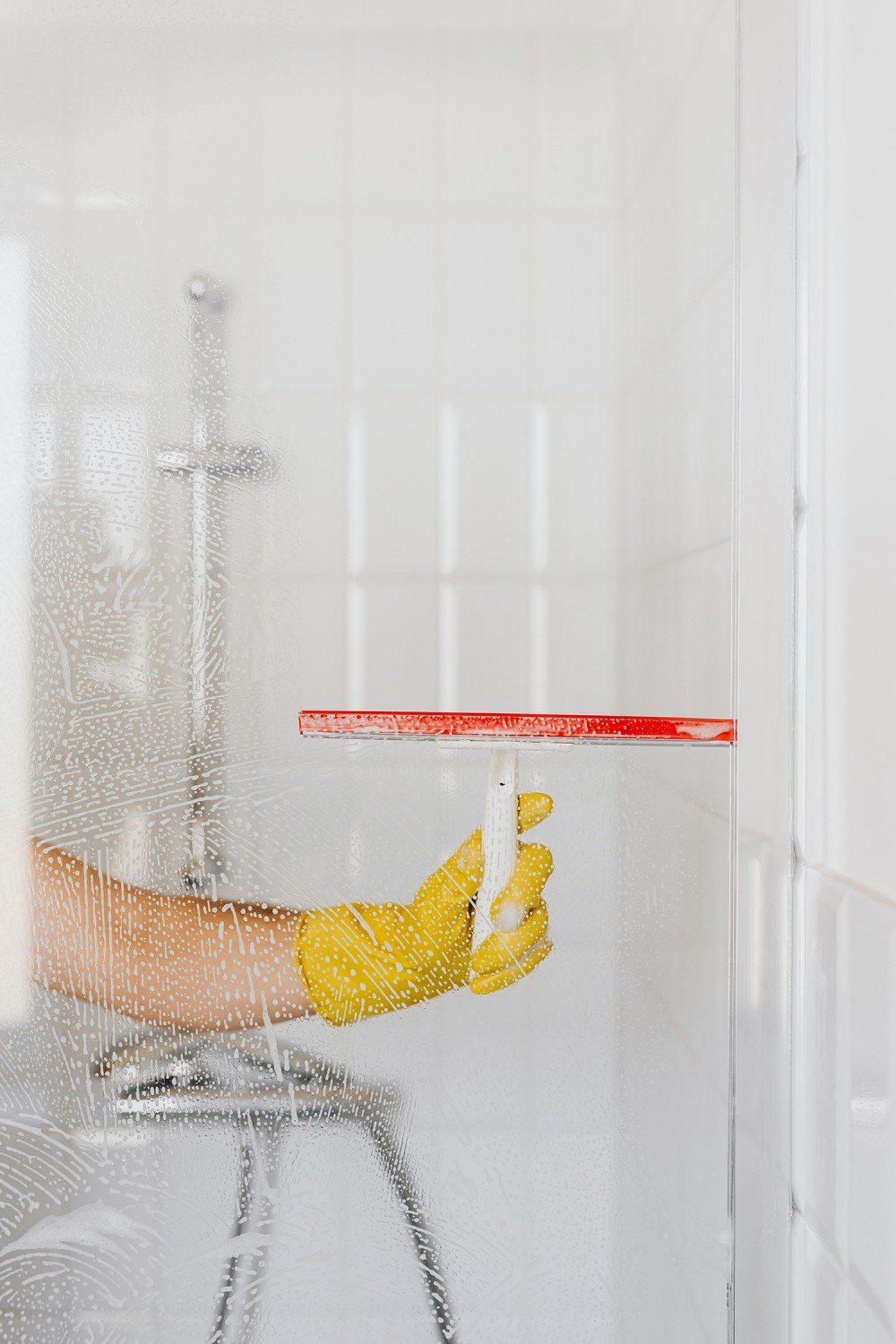 White vinegar helps a woman clean limescale off the glass of a shower enclosure. Wearing gloves, she wipes away the water droplets with a squeegee.