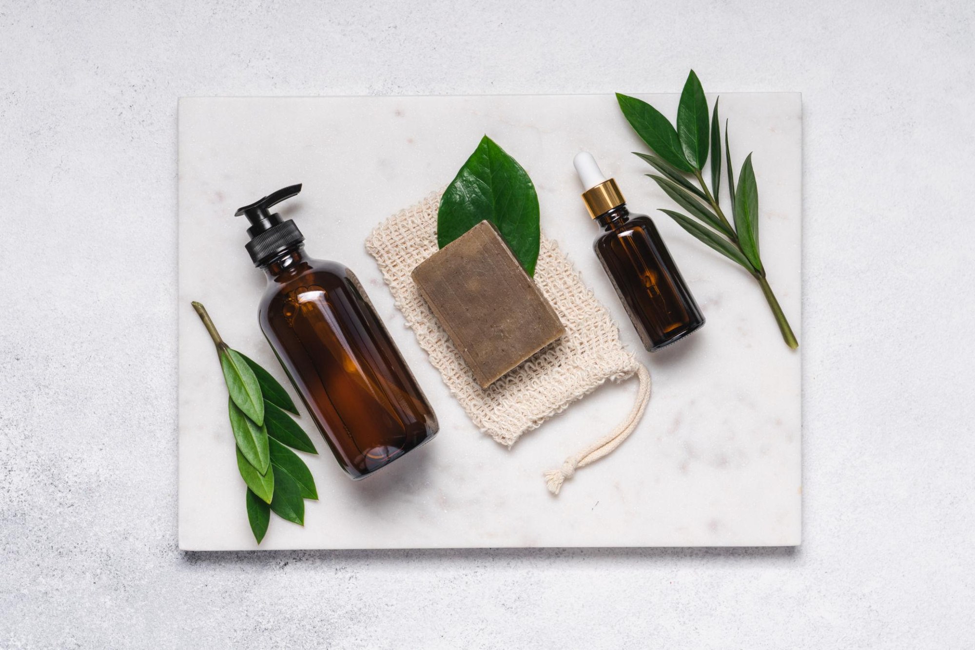 Studio shot of Handymade natural cosmetics — pump dispenser, soap, sisal pouch and dropper on marble.