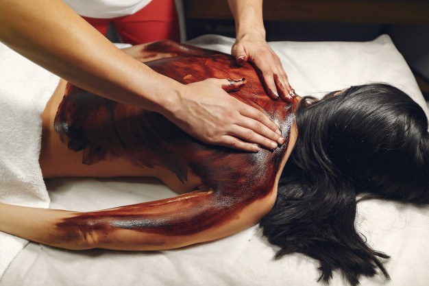 Masque corporel à l’argile cosmétique appliqué sur le dos d’une femme allongée sur une table de massage.