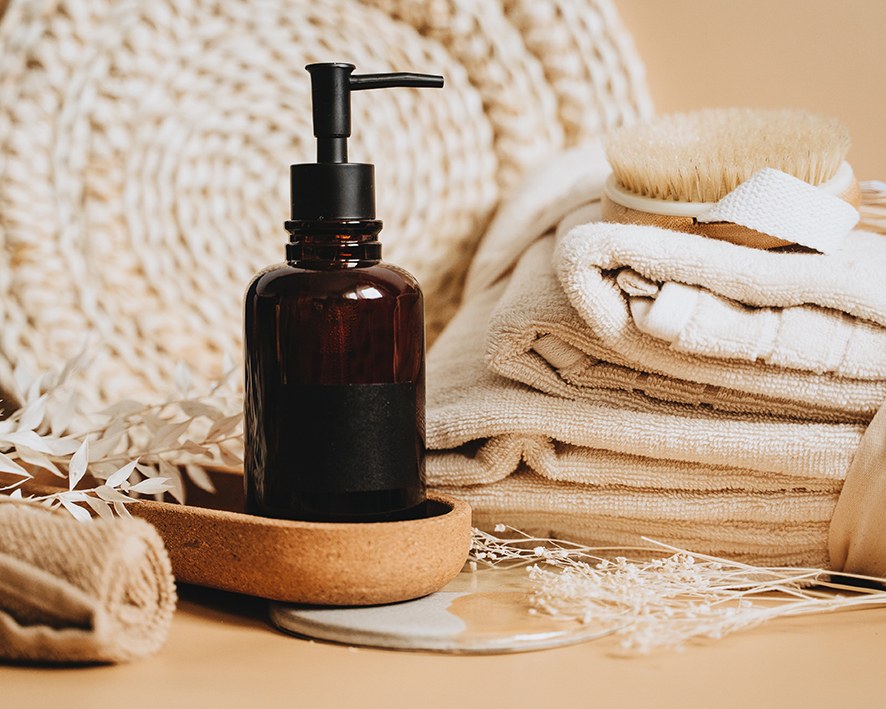 Hausgemachtes Haarshampoo für Einsteiger in einer braunen Glasflasche mit Pumpspender steht auf einem Tisch in einem Stillleben mit Handtüchern, einer Bürste und anderen Waschmitteln.