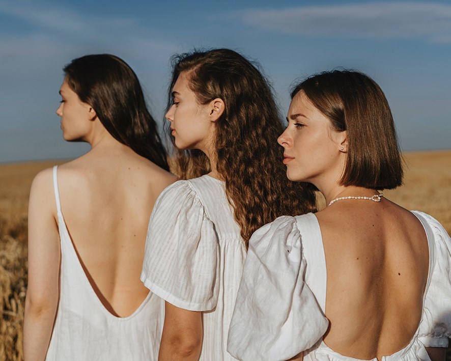 Drei Frauen mit unterschiedlichen Haartypen in weißen Kleidern stehen auf einem Getreidefeld.