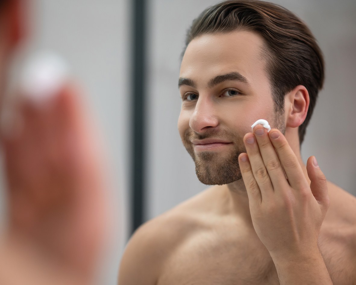 A man stands in front of a mirror and applies face and beard serum to his face.