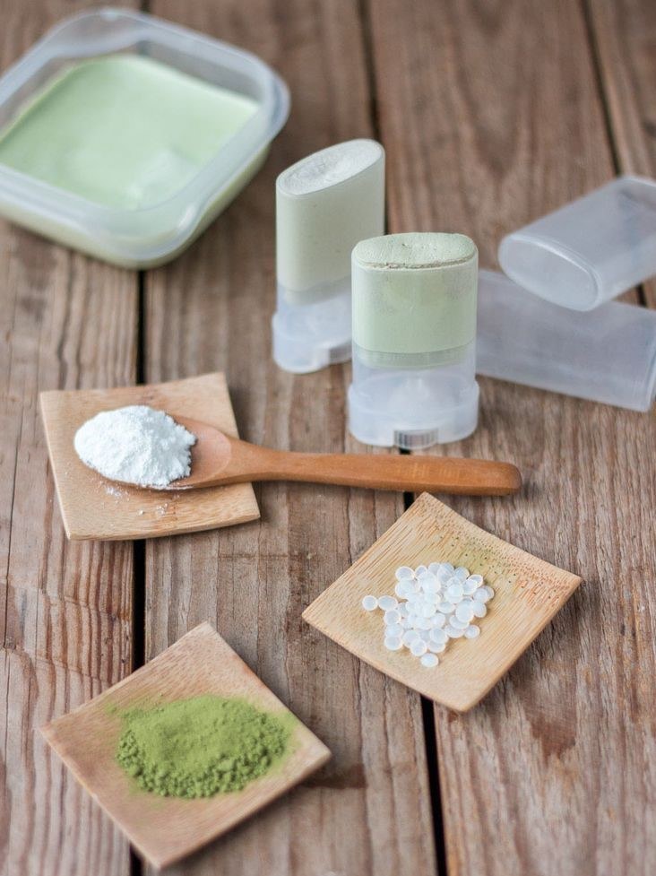 Zwei gefüllte Deodorant-Sticks und eine Dose mit fester Masse auf einem Holztisch, daneben weißes und grünes Pulver sowie Wachspastillen.