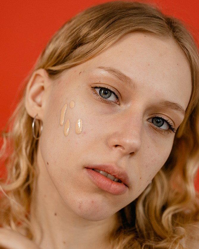 Eine junge Frau hat drei Tropfen flüssige Mineral-Foundation im Gesicht und steht vor einem roten Hintergrund.