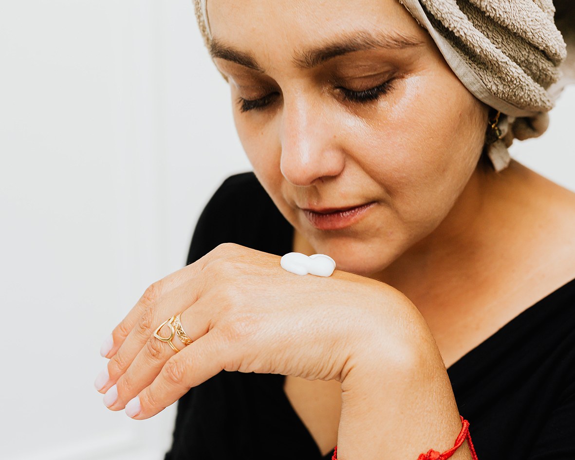 A woman has winter face cream on her hand and leans closer so that she can apply it to her skin. She is in the bathroom and has a towel on her head.